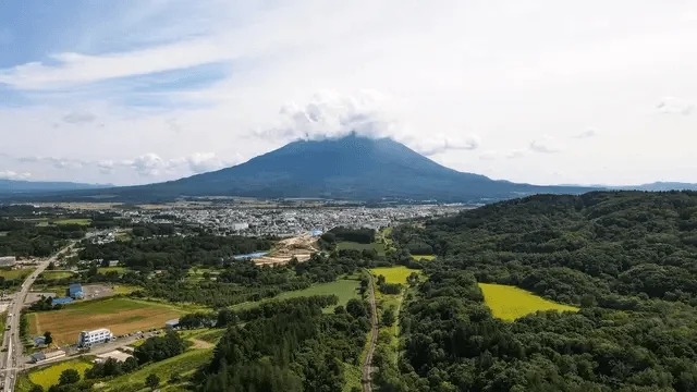 雲に覆われた雄大な山を背景に、広がる街並みと豊かな森林、畑が広がる壮大な風景を捉えた空撮写真。