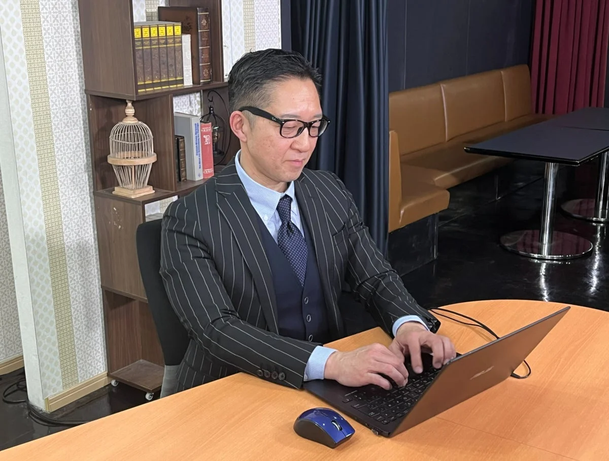 ピンストライプのスーツを着た男性が、木製のデスクでノートパソコンを操作しています。背景には本棚やカフェのような座席があり、ビジネスシーンでの集中した作業風景です。