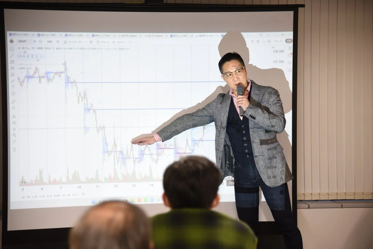 男性がプロジェクターに映し出された金融チャートを指しながら、聴衆に説明しているビジネスセミナーの様子。投資や為替に関する講演を行っている場面と見られる。