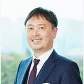紺色のスーツとネクタイを着用したアジア系の男性が、カメラに向かって穏やかに微笑んでいる上半身のポートレート写真です。清潔感があり、プロフェッショナルな印象を与えます。