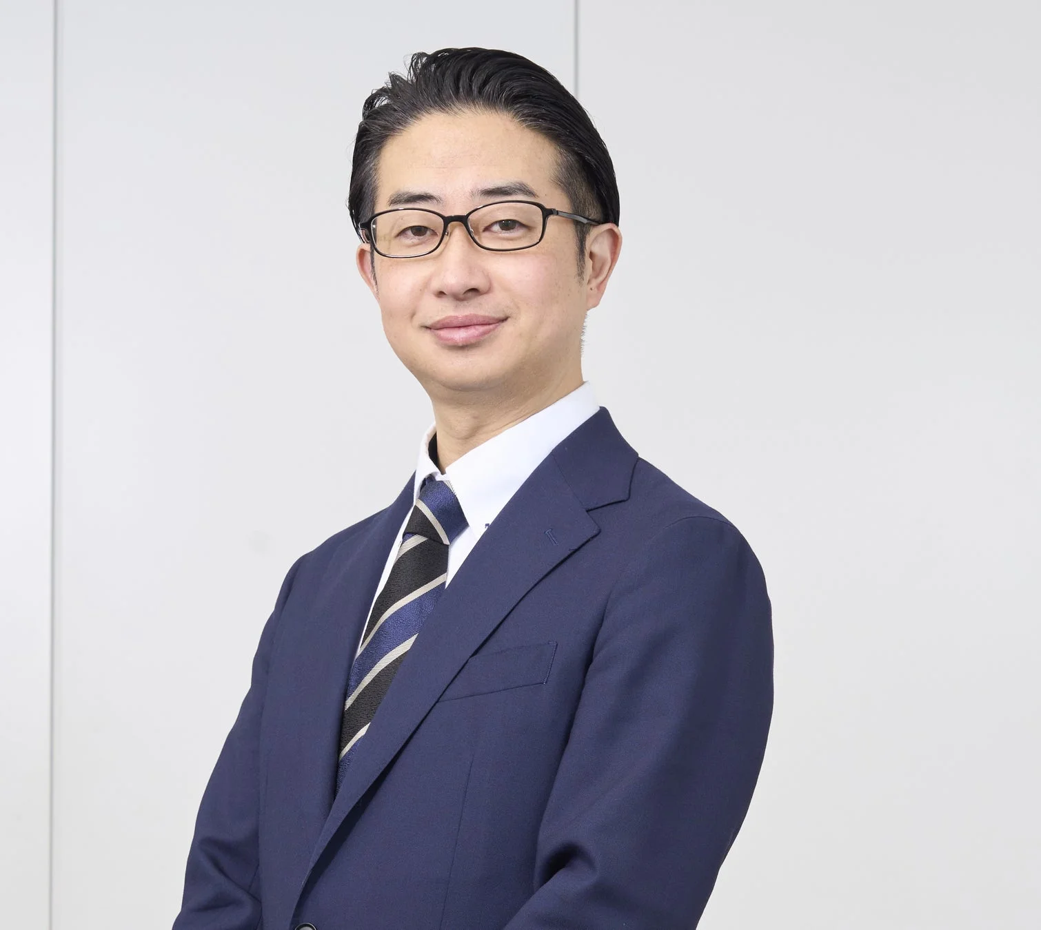 紺色のスーツと眼鏡を着用した日本人男性が、白い背景の前で穏やかに微笑んでいる上半身のポートレートです。